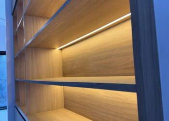 Biblioteca empotrada a medida con estantes iluminados y bauleras superiores para proyecto de arquitectura.